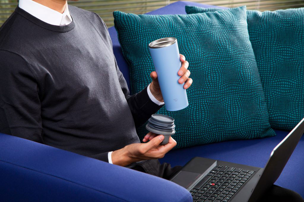 Man sitting on a blue sofa and with a lap top on his lap, and a light blue tumbler in his left hand and the lid with a handle on his right hand.