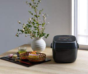 Black rice cooker on the right and a tray with tea and a rice dish to the left, and a round vase with aesthetic branches with leaves
