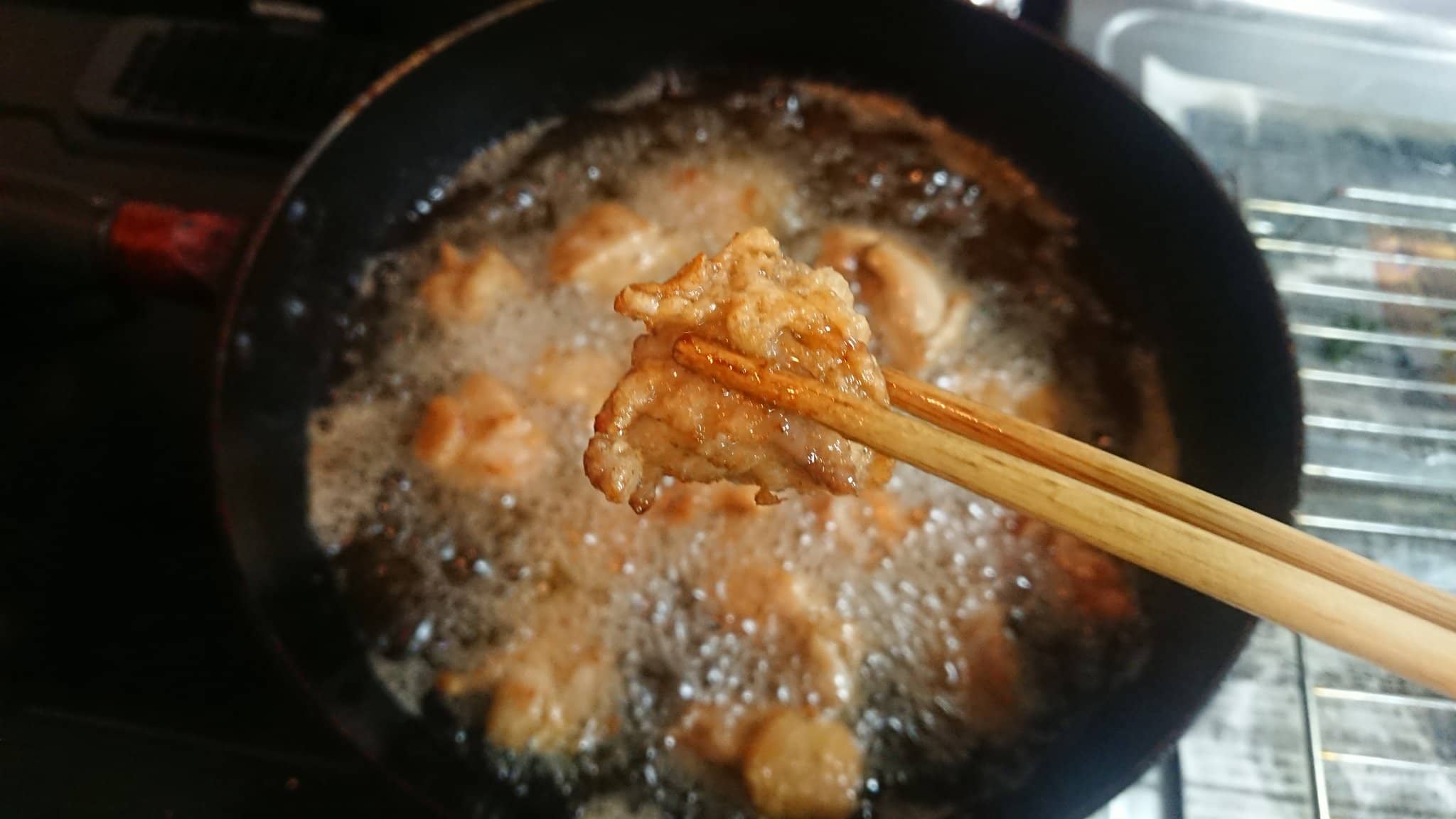 Pan filled with hot bubbling oil and small pieces of chicken and in the foreground a pair of chopsticks holding a piece of fried chicken