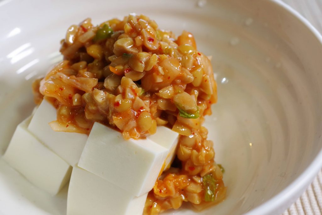 White bowl with cubed tofu topped with chopped natto mixed with spicy sauce and onions.