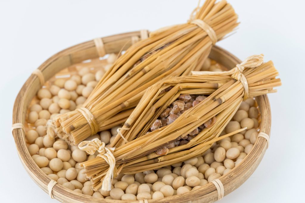 Woven basket filled with soy beans and on top two straw bundles tied on each end filled with fermented soy beans.