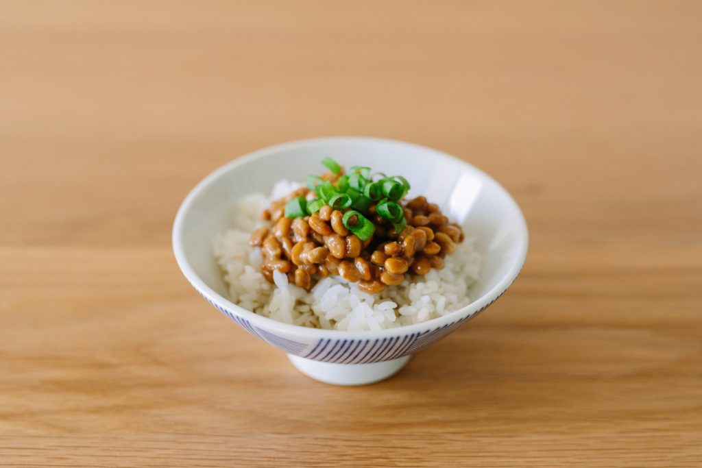 Wooden table with a small bowl with blue stripes on the outside filled with white rice, topped with natto and chopped scallions.