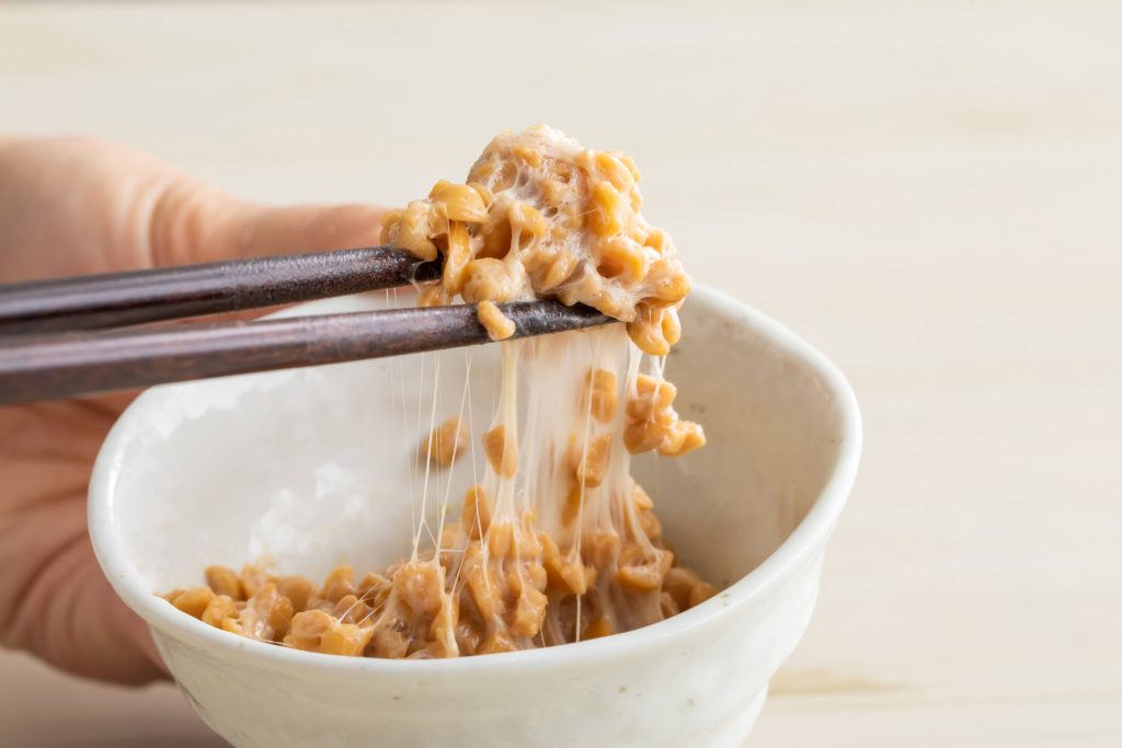 Hand holding a bowl filled with fermented soy beans and they are taking some out with their chopsticks. There are visible strings as they are taking a portion.