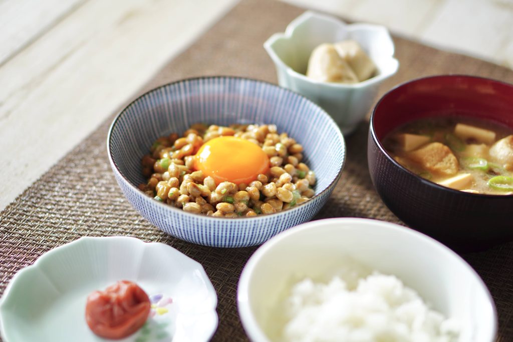 Traditional Japanese breakfast set on a table with small bowls filled with rice, miso soup, pickles, and natto topped with an egg. 