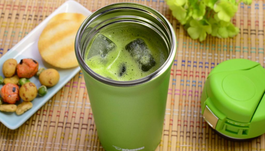 Stainless mug filled with iced matcha beverage accompanied with a white plate filled with rice crackers.