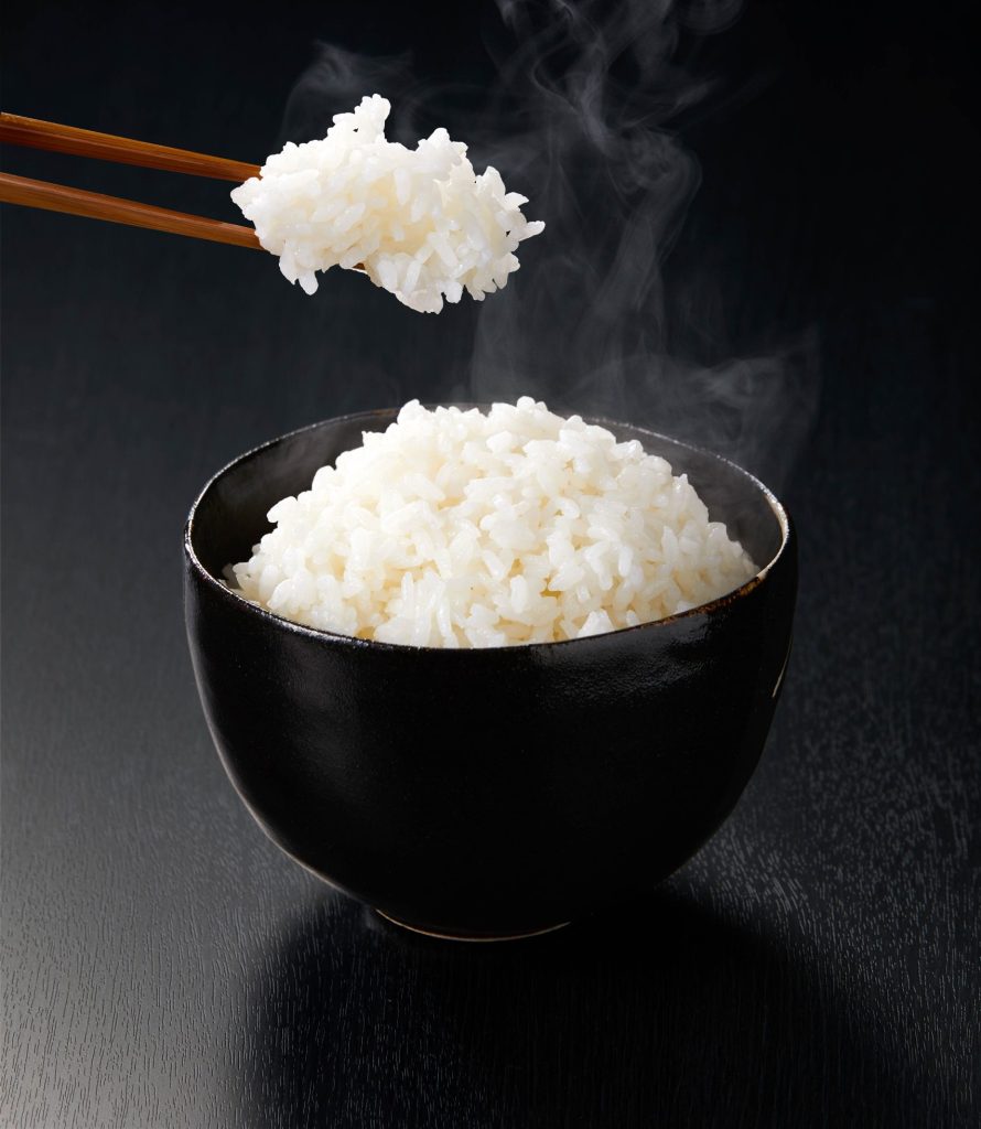 Freshly cooked rice in a traditional Japanese bowl and a pair of chopsticks holding a bite of rice with steam rising