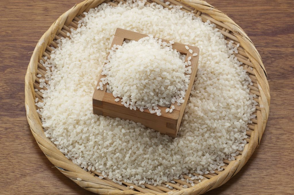 Uncooked white short grain rice in a flat woven basket, in the center a wooden square container filled with rice