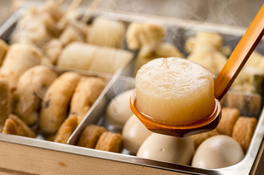 Wooden tray with internal metal compartments filled with broth and oden ingredients like fish cakes, eggs, and daikon; a wooden ladle in the foreground with a daikon round