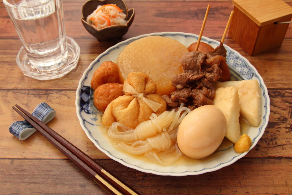 Wooden table in the background with a deep plate filled with oden like an egg, konyaku noodles, fish cake and radish; served with a side of pickles and a glass of water.