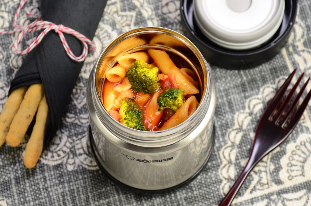 Stainless steel food jar filled with a pasta dish with broccoli and a side of breadsticks on the side