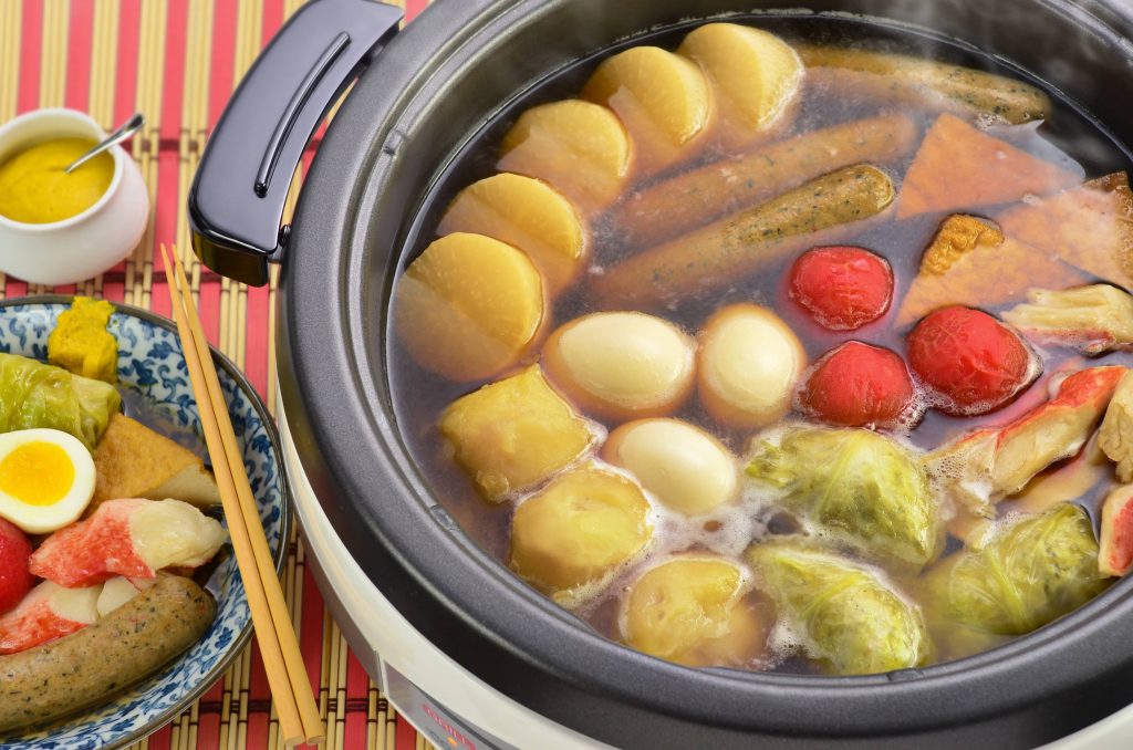 Zojirushi electric skillet filled with hot oden and a plate served on the side with a pair of chopsticks and mustard dipping sauce