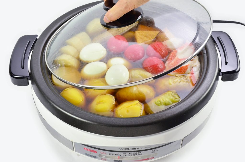Zojirushi electric skilled filled with broth and various oden ingredients like fish cake, sausage, daikon, tomatoes, eggs, and stuffed cabbage.