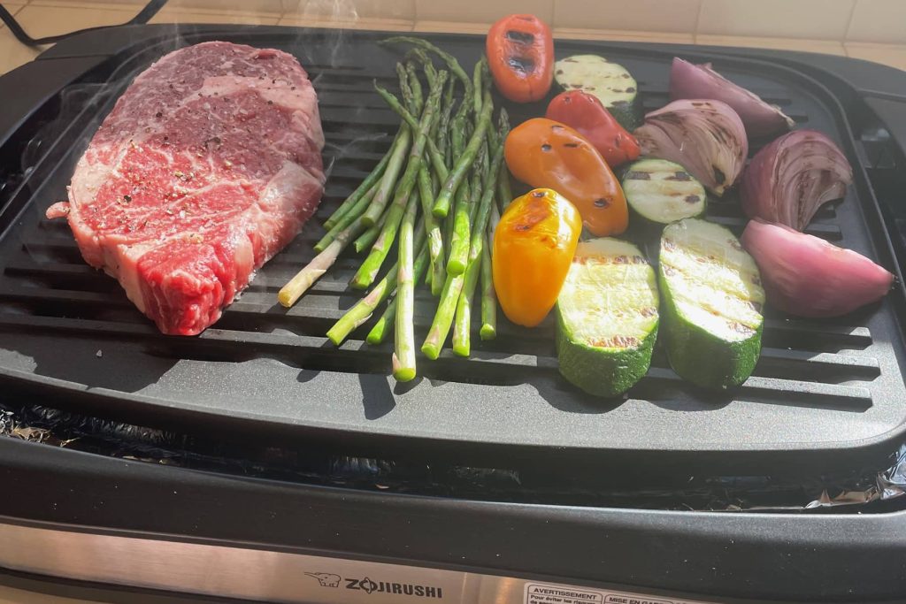 Grilling piece of steak on the electric grill along with asparagus, peppers, zucchini and red onion