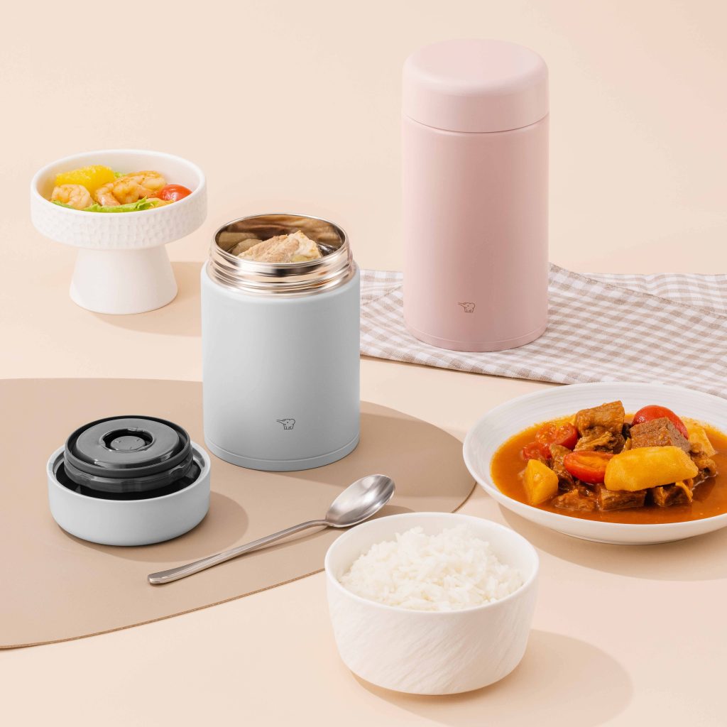 Lunch spread on a light brown table: a large pink food jar in the background and a light blue medium food jar in front, open filled with food, a side of stew, a bowl of rice and veggies