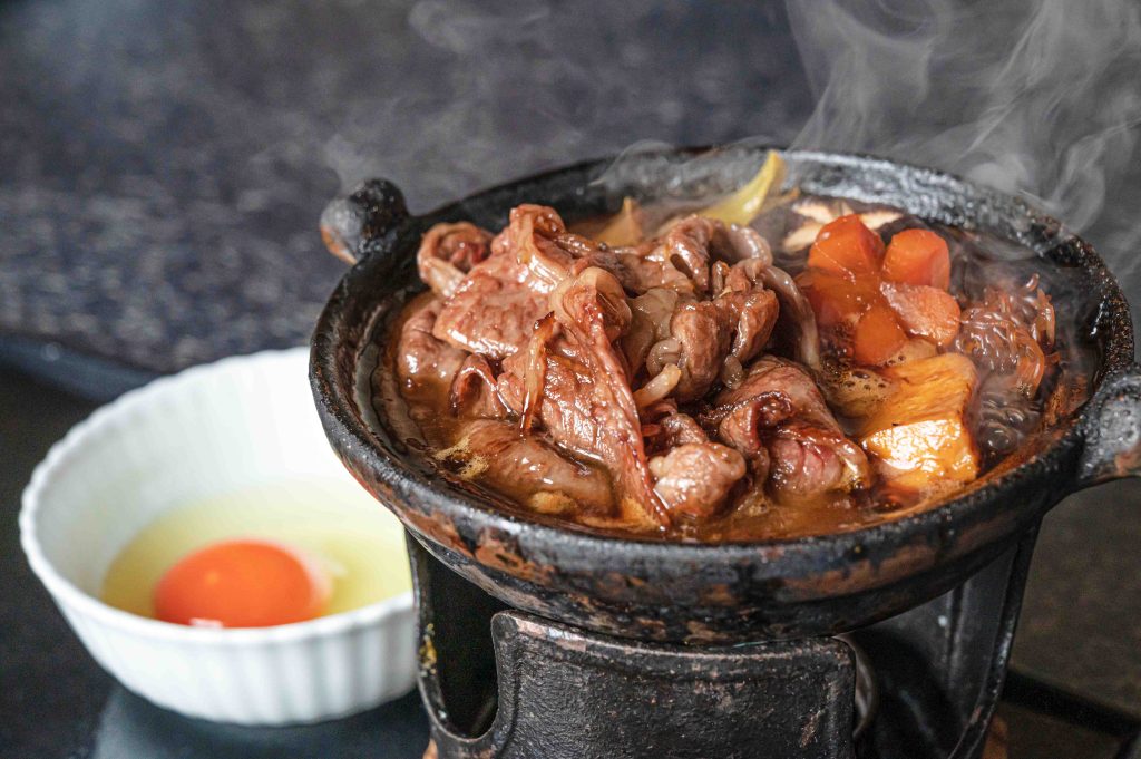 Small iron pan over heat cooking up a meat and veggie stew with a small white bowl with a cracked egg