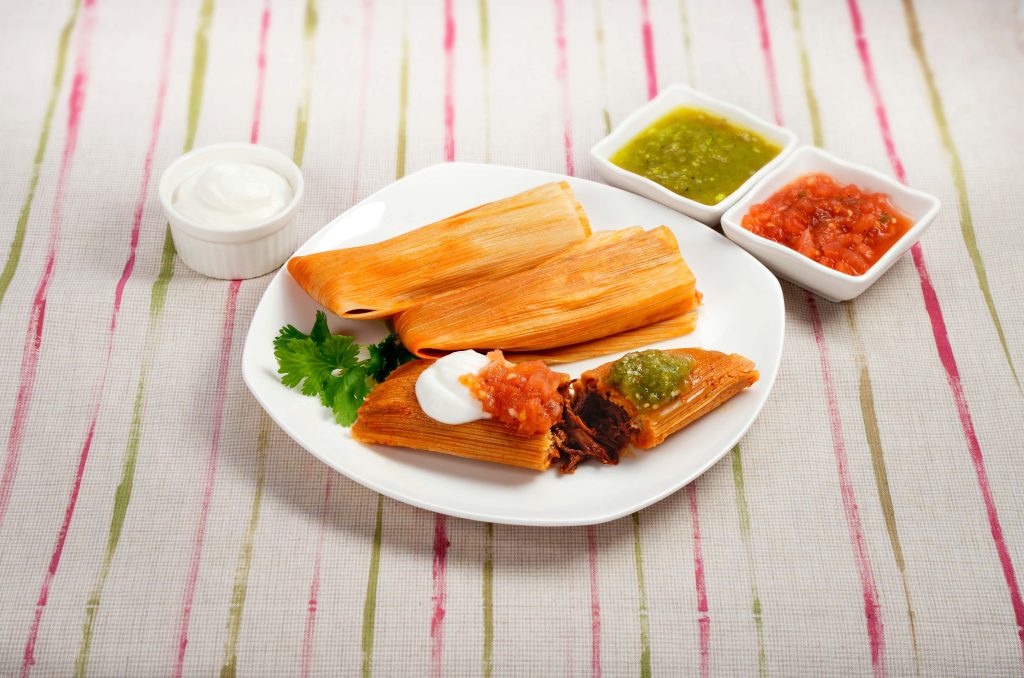 Plate with three beef tamales - one is cut in half topped with green and red salsa, and sour cream. Sides dishes with green and red salsa, and sour cream on the side.