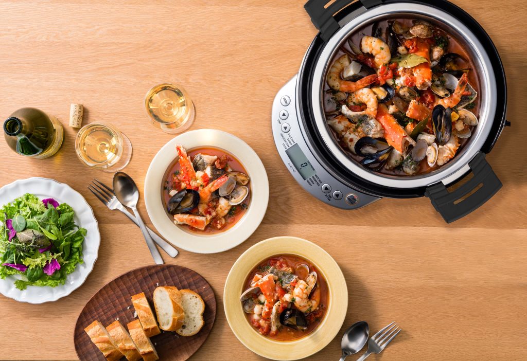 Top view of the multicooker with seafood stew. Besides it two served plates with a side of bread and salad. 