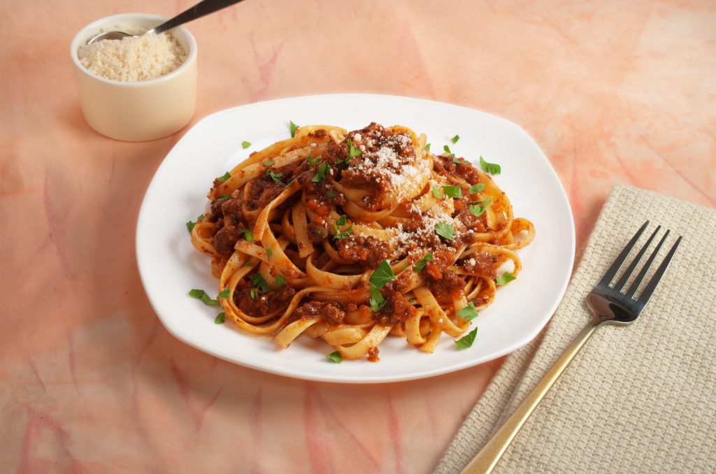 A white plate with a serving of fettuccine bolognese sprinkled with basil and parmesan cheese with a side dish with parmesan cheese on the side. 