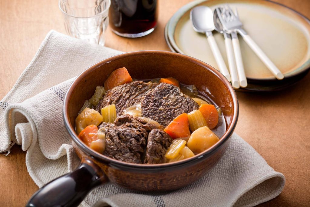 A serving of pot roast with chunks of meat, potatoes and carrots. A plate with four spoons on the side.