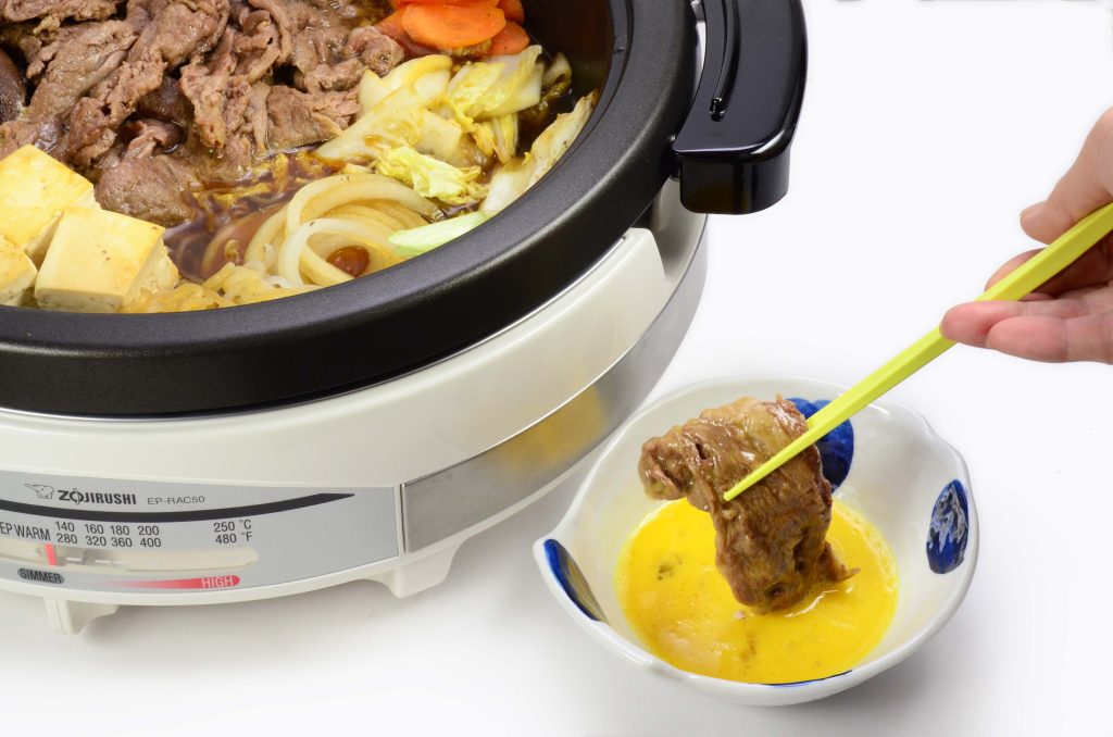 Electric skillet filled with sukiyaki and a small dish with an egg and a person's hand dipping some meat into it.