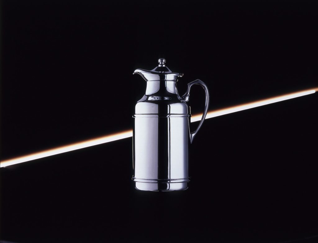 Chrome carafe in the center of a black background with a white light in a diagonal line in the back