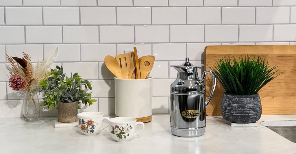 Pelican pot on a kitchen counter with a plan to the right and wooden kitchen utensils and two vintage cups with a flower design on the left.