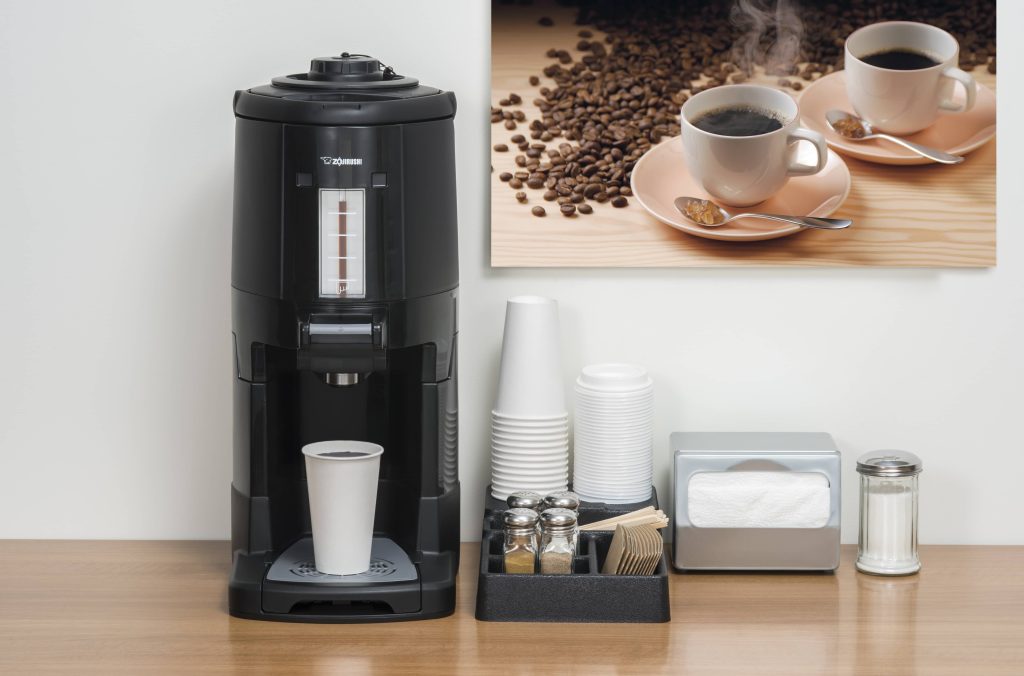 Gravity pot with coffee cup filled with coffee under it's spout and a condiment and paper cup organizer, silver napkin dispenser and sugar jar to the right, above on the wall is an image of two hot coffee cups with beans spilled over the table. 