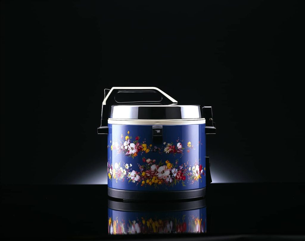 The royal blue Hanagumo rice cooker with a flower design around it and stainless trimming over a black reflective surface and a black background