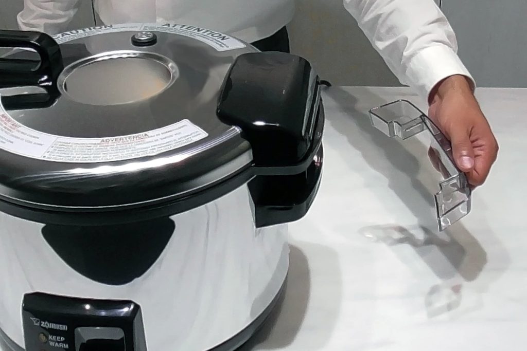 Raised view of the rice cooker with a person removing the transparent dew collector on the right side 