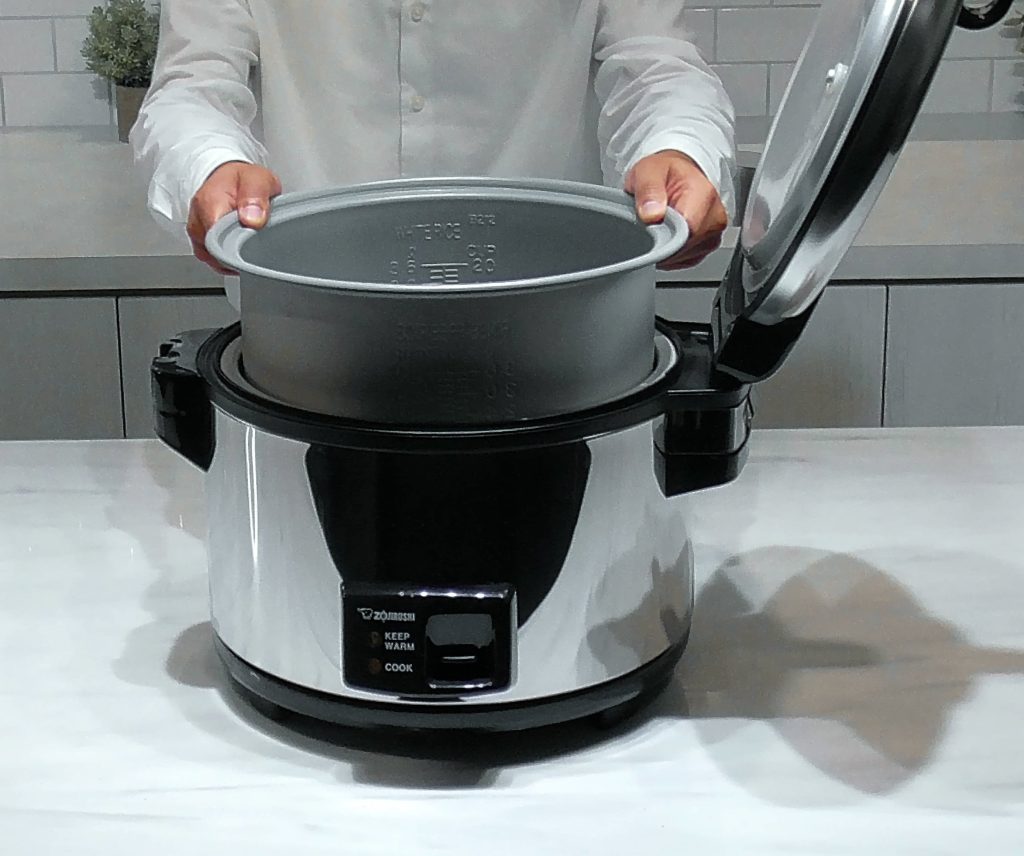 Large stainless rice cooker with the lid open and a person in the background lifting out the cooking pan