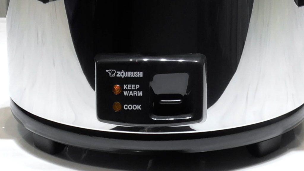 Close up of the bottom front of the rice cooker with a black panel with a switch that has keep warm and cook setting with a small light bulb on the side of each setting