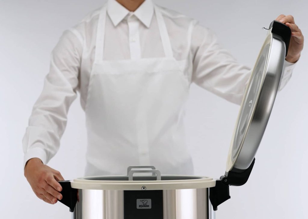 A person wearing a white shirt and apron opens the lid of the rice warmer