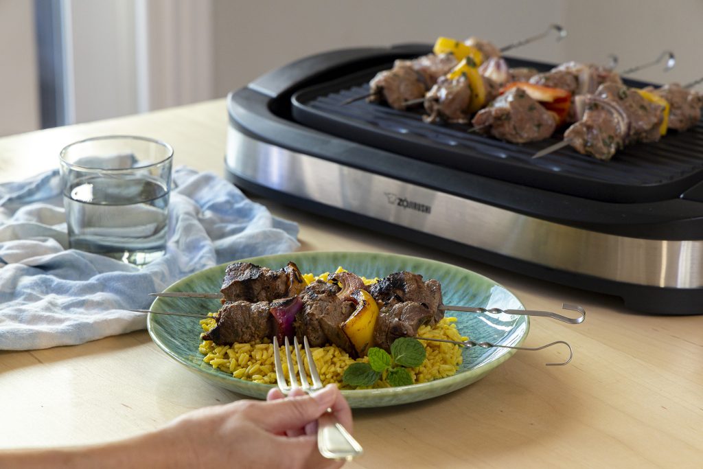 Lamb Kebabs with Yellow Rice on a plate with lamb kebobs being grilled on an indoor grill in the back.