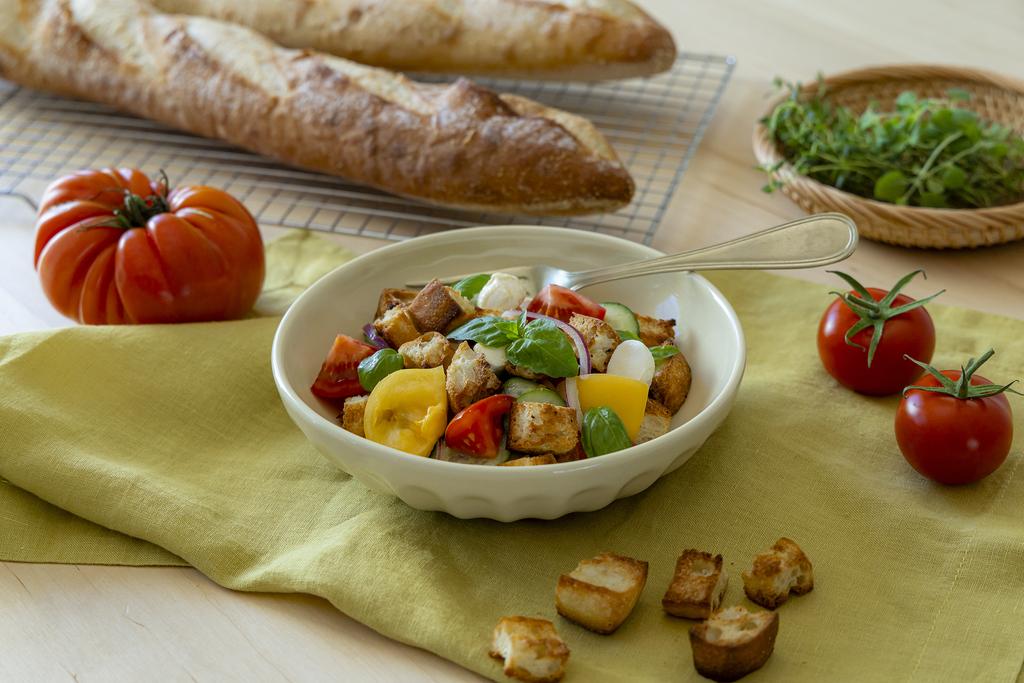 Panzanella is in a white bowl, tomatoes and baguette around the table
