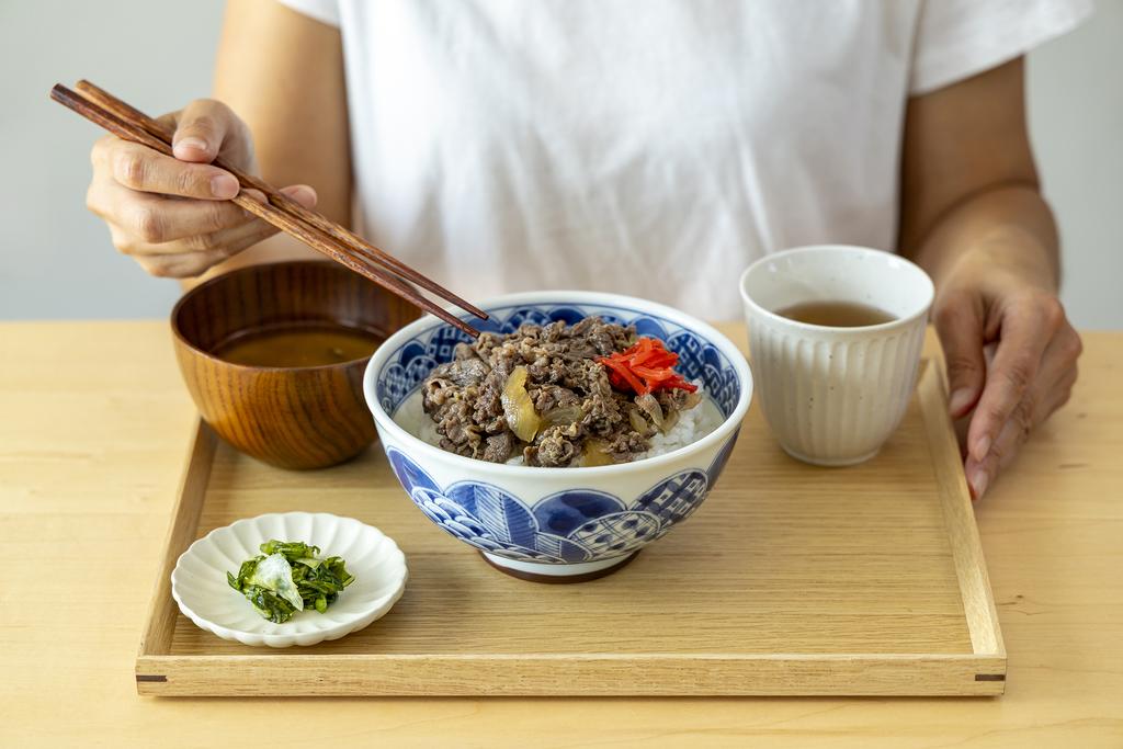 Gyudon is on the table next to miso soup, tea, and garnish, a person is trying to eat with chopsticks on their hand