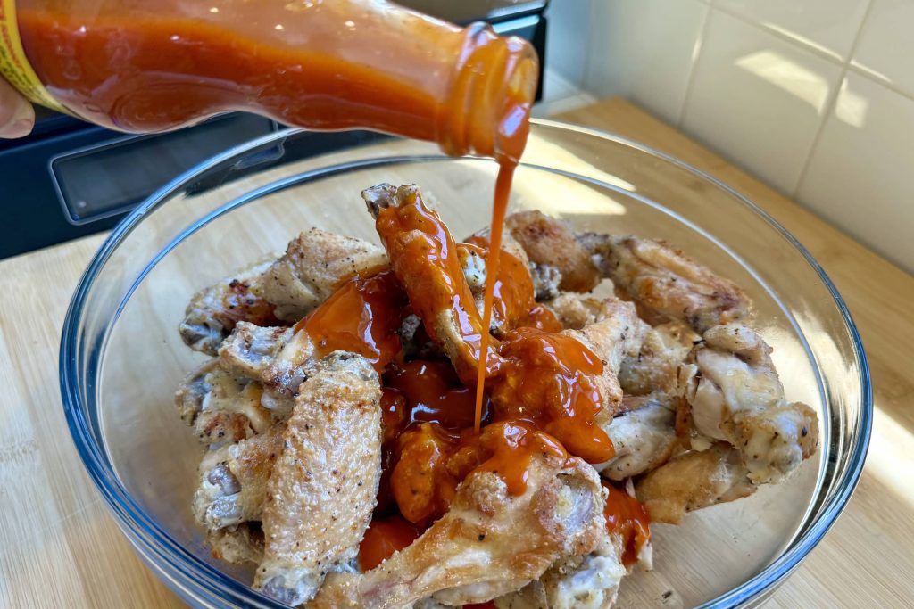 Pouring hot red sauce on bowl of baked chicken wings