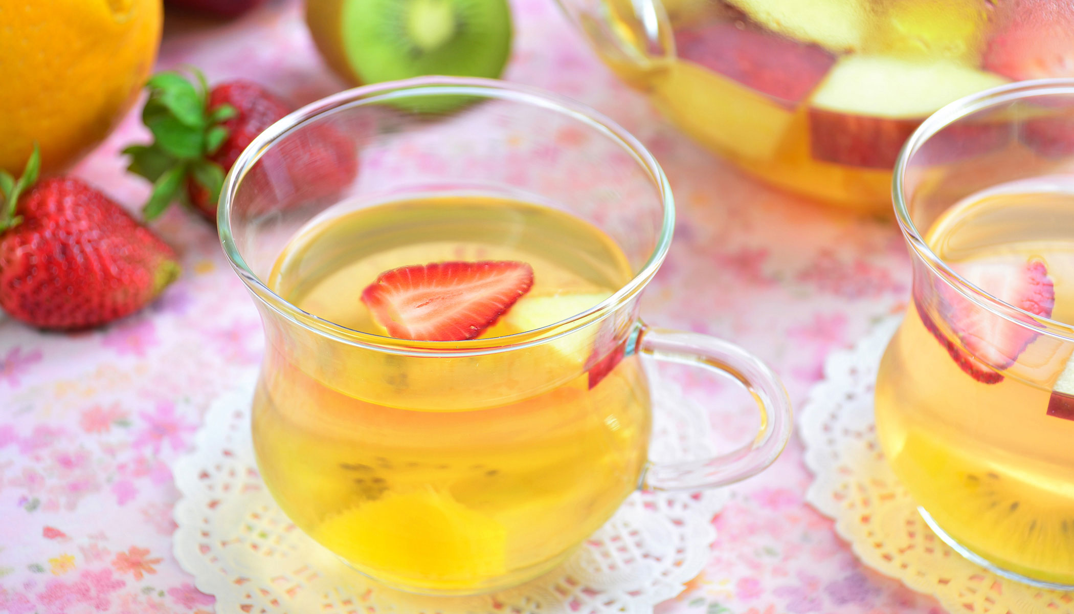 Two cup of tea with kiwi and strawberry slices with strawberries, kiwi, and an orange in the background. There is also a large pitcher with tea and fruits.