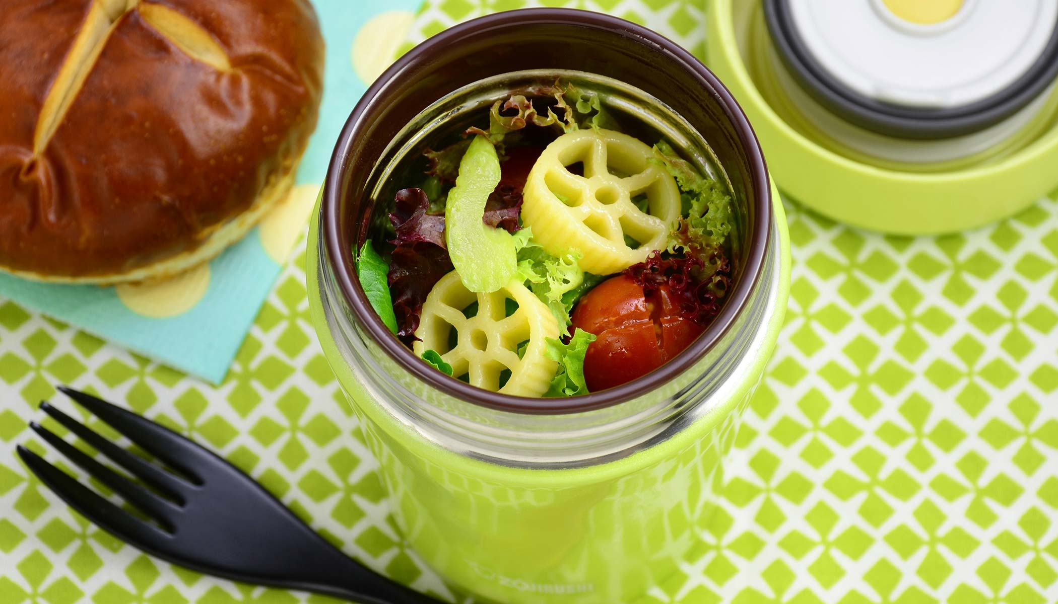 Pinwheel pasta salad with lettuce and tomato served in a green vacuum insulated food jar and a pretzel bun