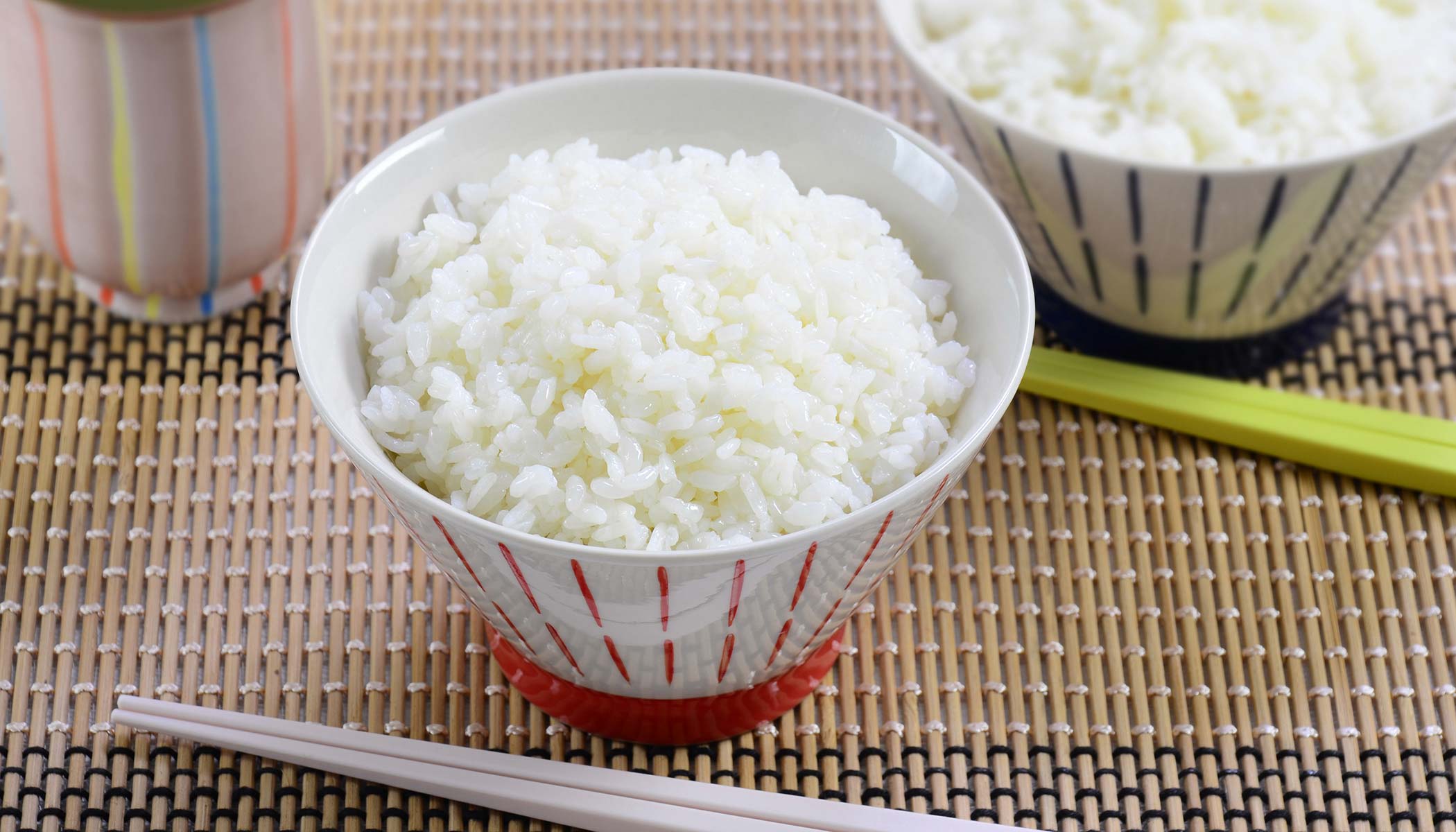 White bowl with a red bottom and red lines from bottom to top, filled with white rice and a pair of chopsticks in front. Another bowl of rice in the back and a cup of tea