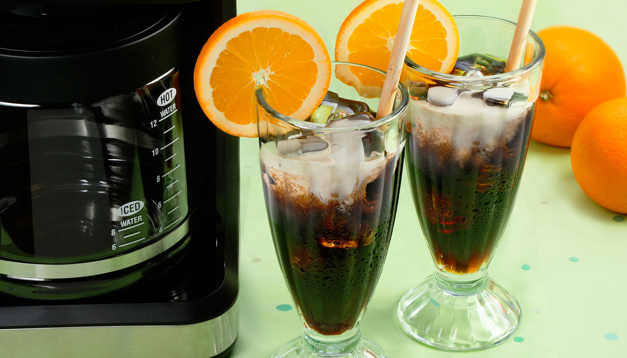 Two glasses of iced coffee with a splash of cream topped with an decorative orange slice and a straw. Two oranges in the background and a coffee maker on the left.